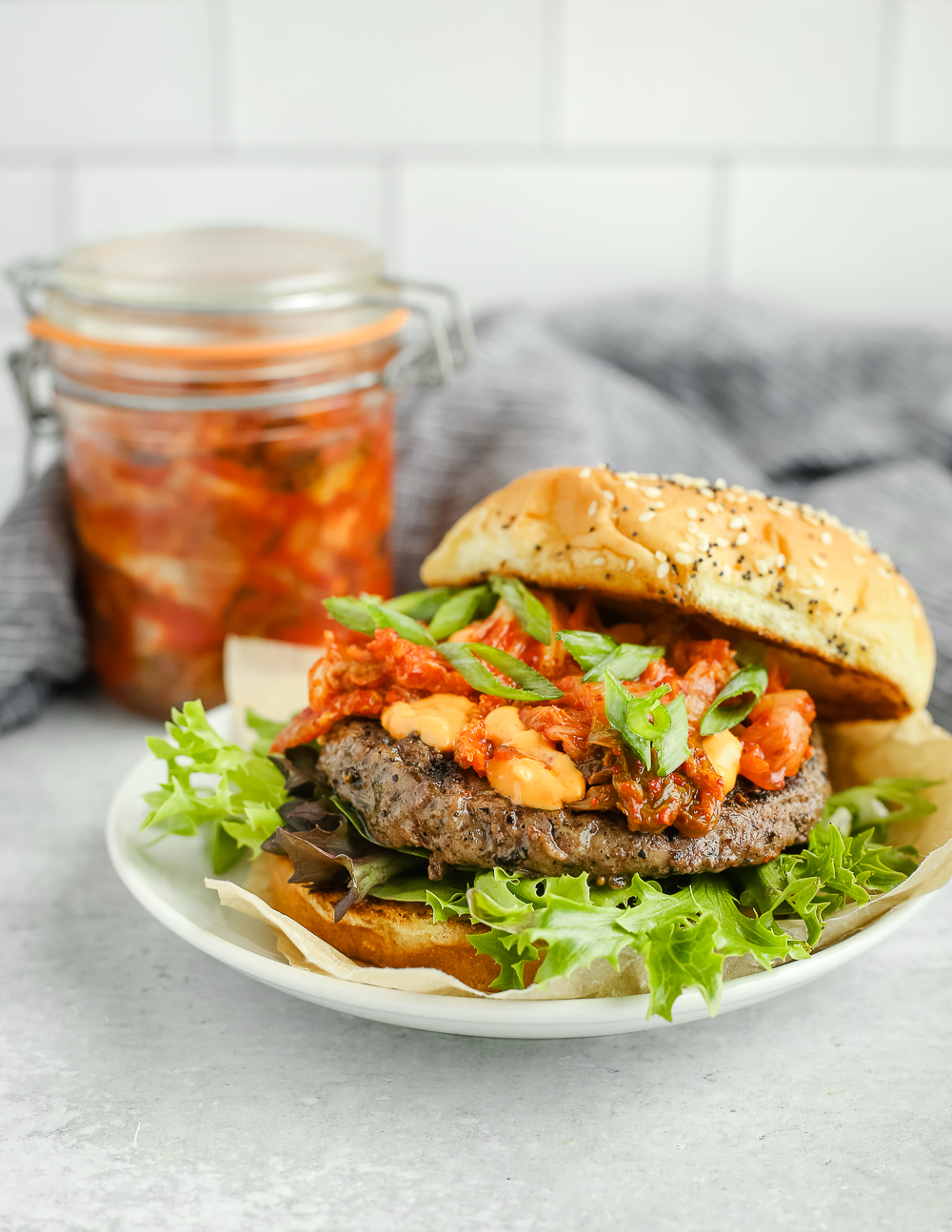 A side view of a beef burger topped with spicy mayo and kimchi with lettuce and green onion slices, served on a small white dish with a jar of prepared kimchi in the background