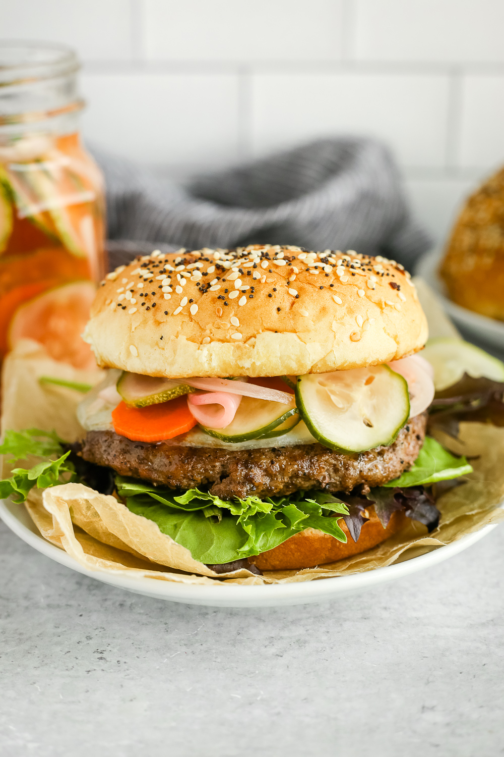 Side view of a cheeseburger topped with colorful veggies, served on a small white dish with a jar of pickles and toasted buns in the background