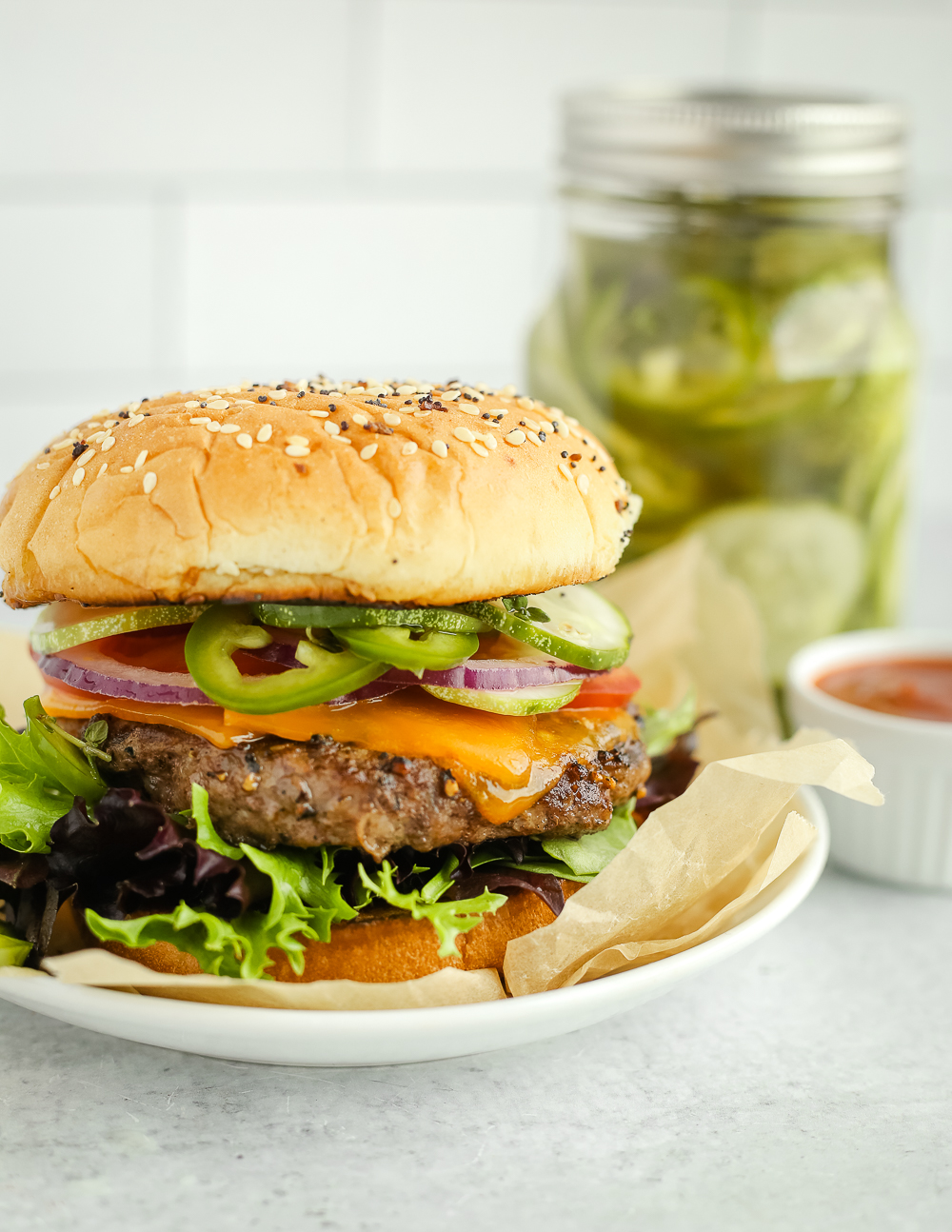 Side view of a classic beef burger on a toasted bun with a pickle pairing of cucumbers, jalapeños, and summer herbs, served on a small white dish with the jar of pickles in the background