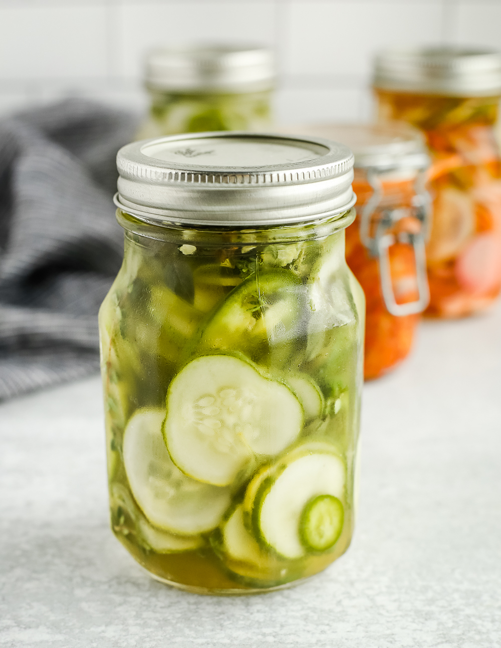 A clear glass jar filled with quick pickled cucumbers and onions with a silver lid, with other jars of various types of quick pickles in the background