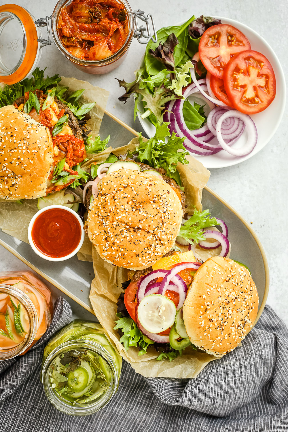 Overhead view of a burger platter with three types of beef burgers, topped with various quick pickle pairings, with side dishes and condiments scatter throughout the frame