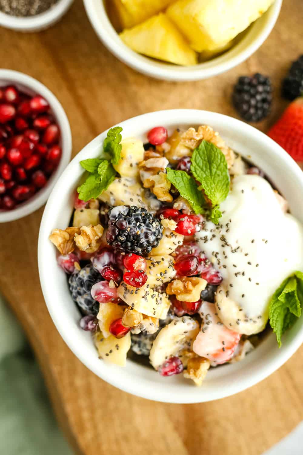 Overhead view of a fresh fruit salad surrounded by pineapple, pomegranate, and fresh berries