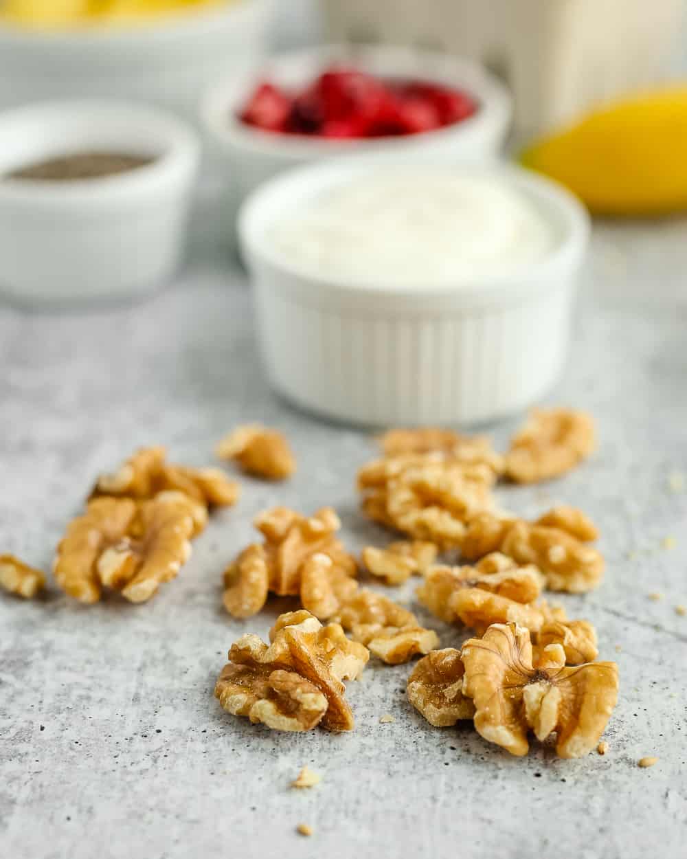 A handful of shelled walnuts on a grey kitchen countertop with white ramekins of other ingredients in the background