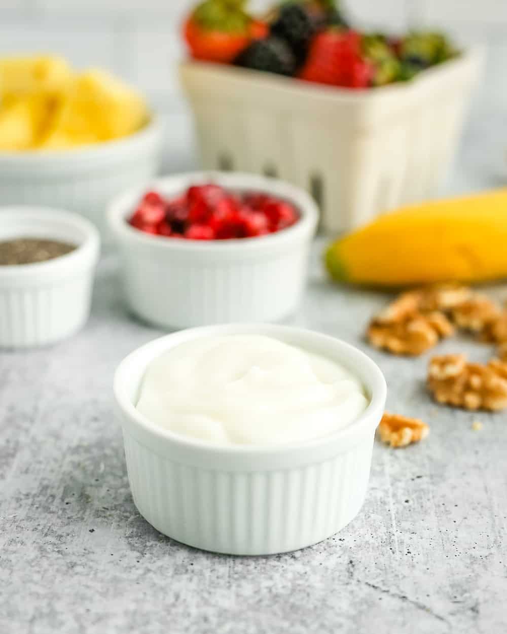 A small white ramekin of vanilla yogurt on a kitchen countertop surrounded by other fresh fruit salad ingredients