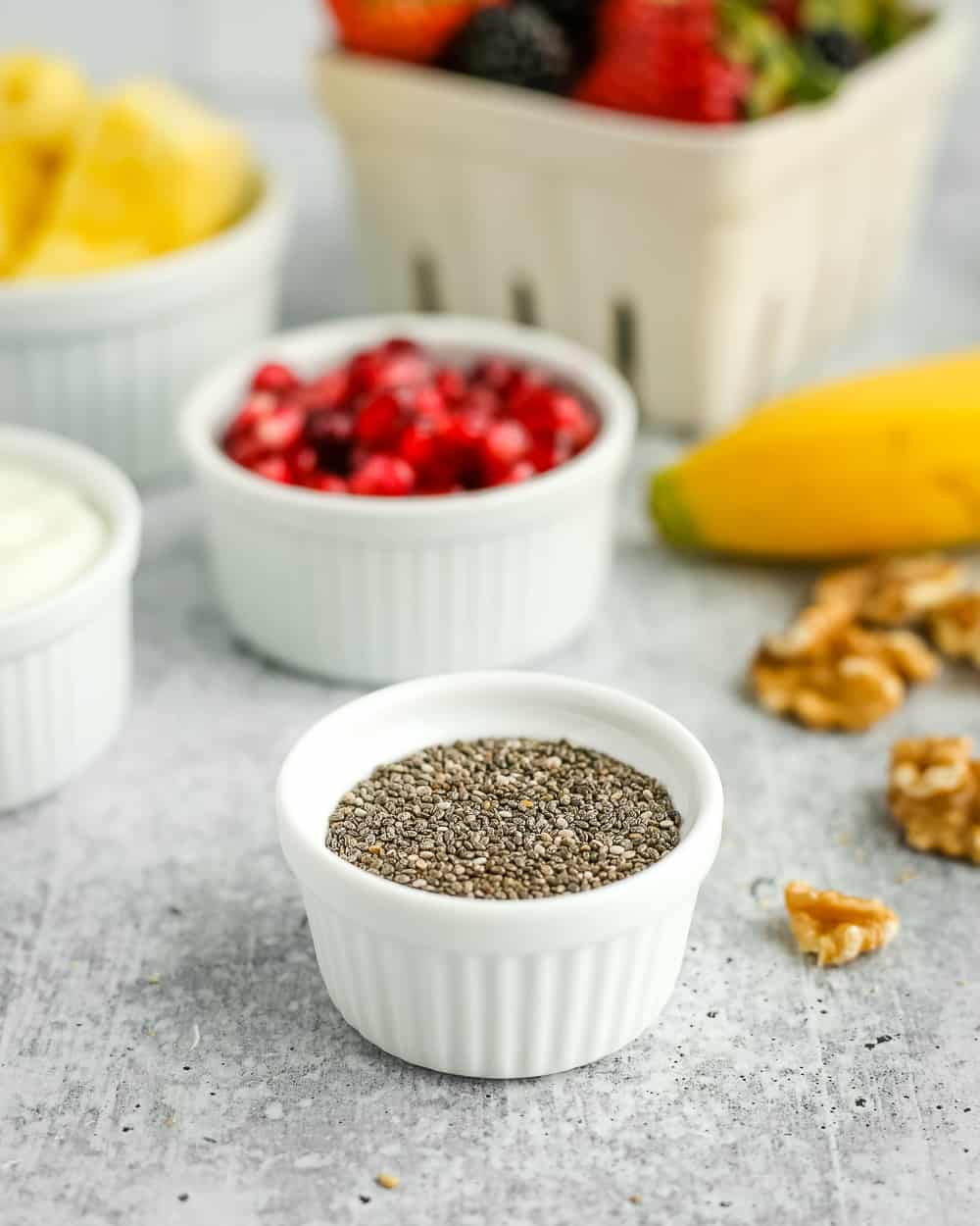 A small white ramekin of chia seeds with pomegranate, an unpeeled banana, fresh berries, chopped pineapple, and walnuts in the background
