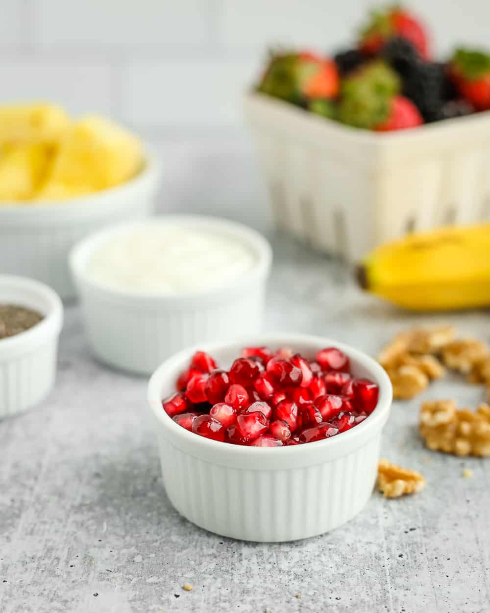 A small white ramekin filled with fresh pomegranate arils with other fresh fruit salad ingredients in the background
