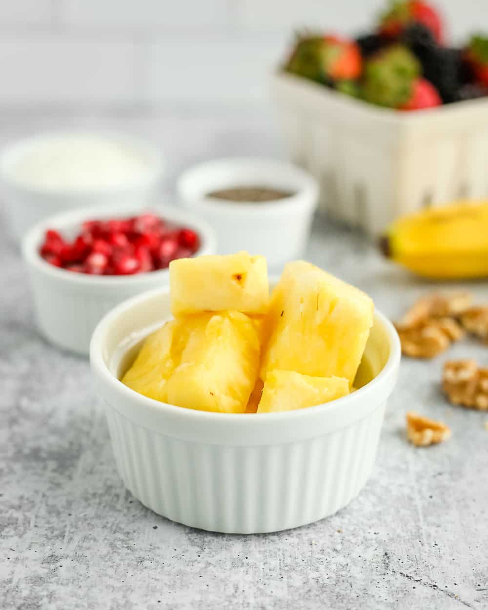 A small white ramekin with chopped pineapple chunks with other fresh fruit salad ingredients in the background