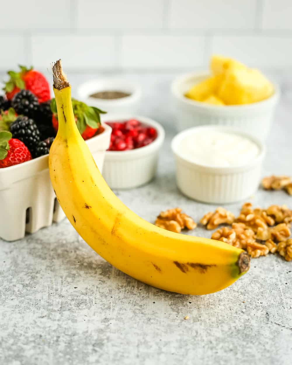 An unpeeled banana on a kitchen counter, propped up by a container of fresh berries and surrounded by other fruit salad ingredients