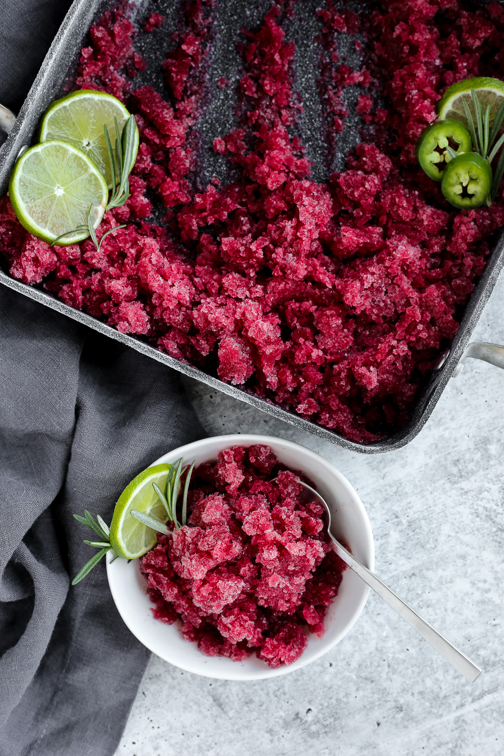 Overhead view of a shallow baking dish with cranberry granita with a small white dish of a single serving garnished with lime and fresh rosemary