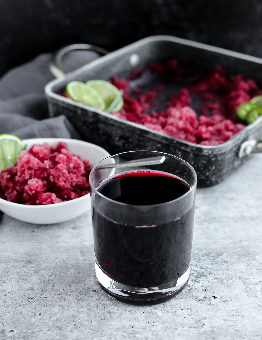 A glass of 100% cranberry juice in front of a white dish with frozen cranberry jalapeño granita and limes