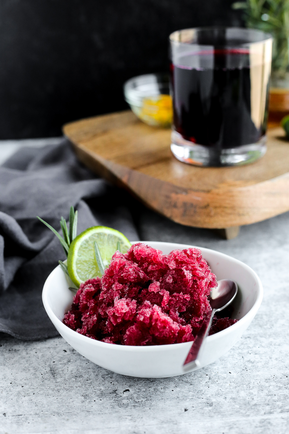 A small white dish with cranberry granita, garnished with sliced lime and a silver spoon, sits in front of a serving tray with a class of cranberry juice