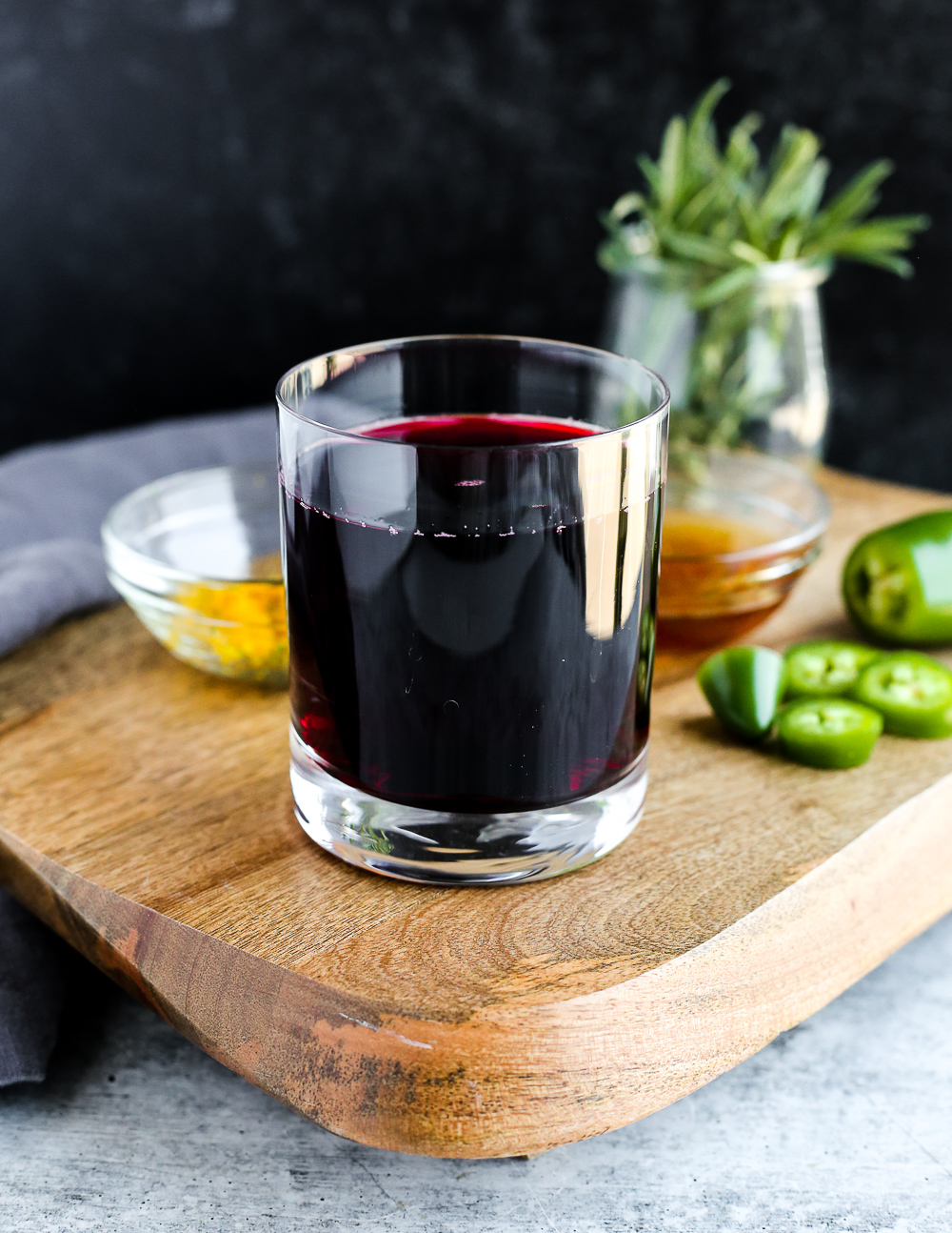 A cocktail glass of 100% cranberry juice on a serving tray, surrounded by ramekins of honey, sliced jalapeños, orange zest, and fresh rosemary sprigs