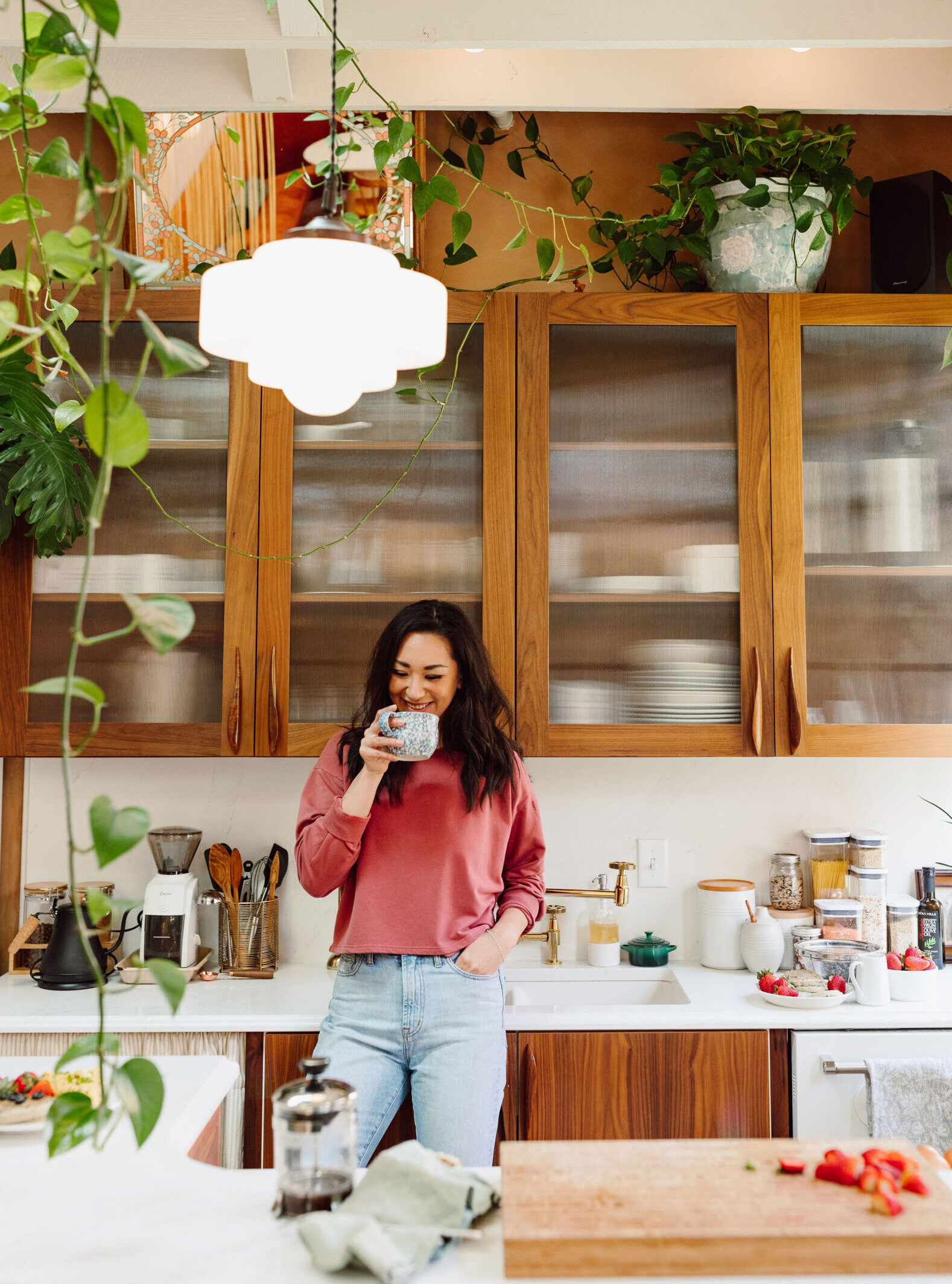 A smiling woman stands in a kitchen holding a mug of coffee, looking content and happy before eating breakfast.