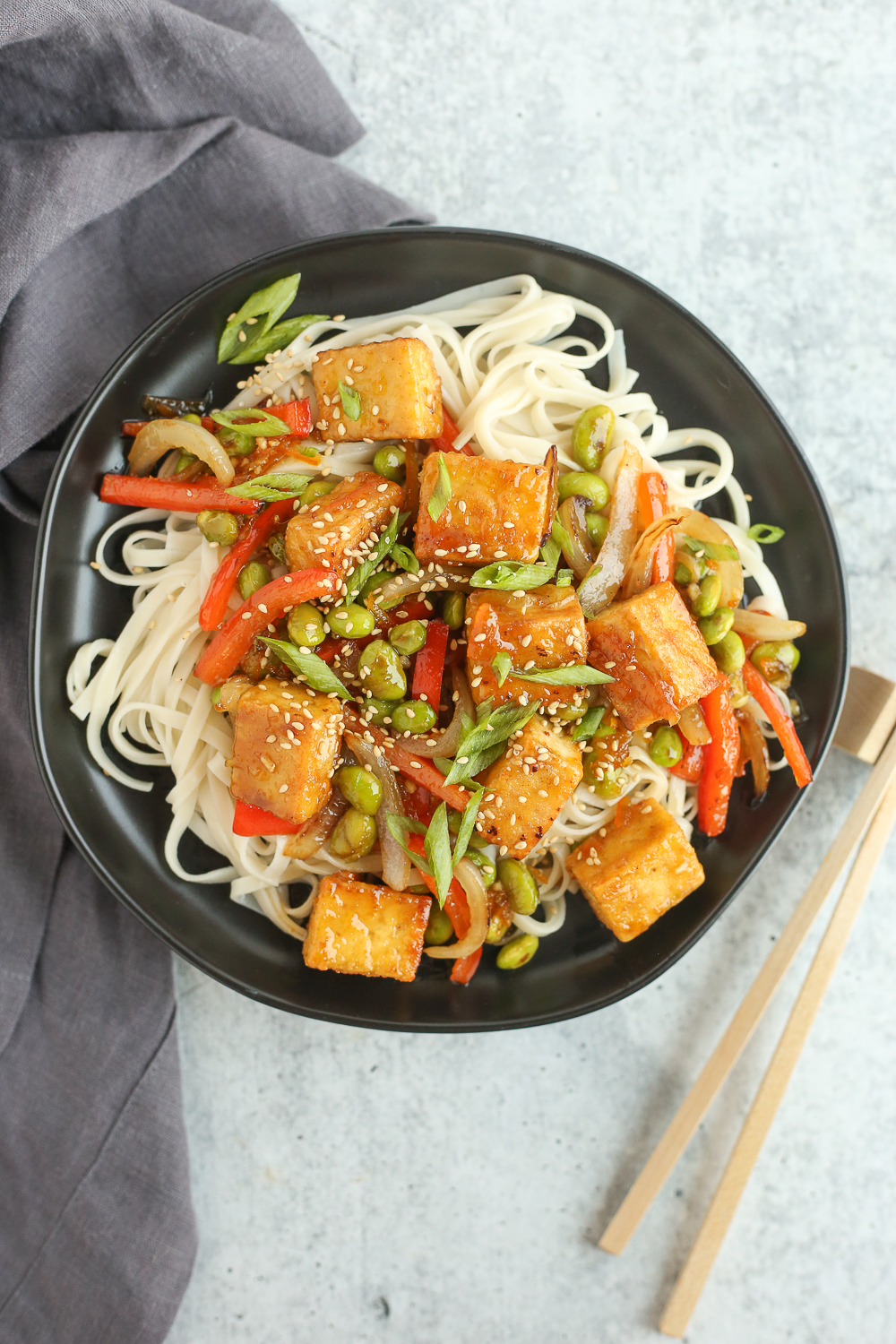 An overhead view of a vegetarian stir fry with crispy orange tofu with noodles, served on a black ceramic plate with chopsticks