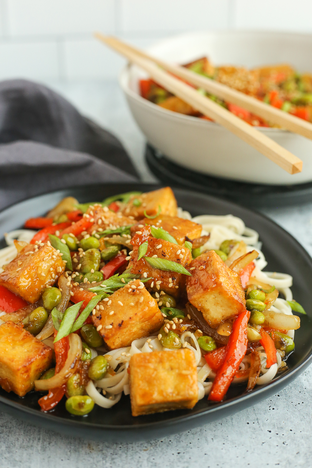 A crispy orange tofu and vegetarian stir fry served with noodles on a black plate with a bowl and chopsticks in the background