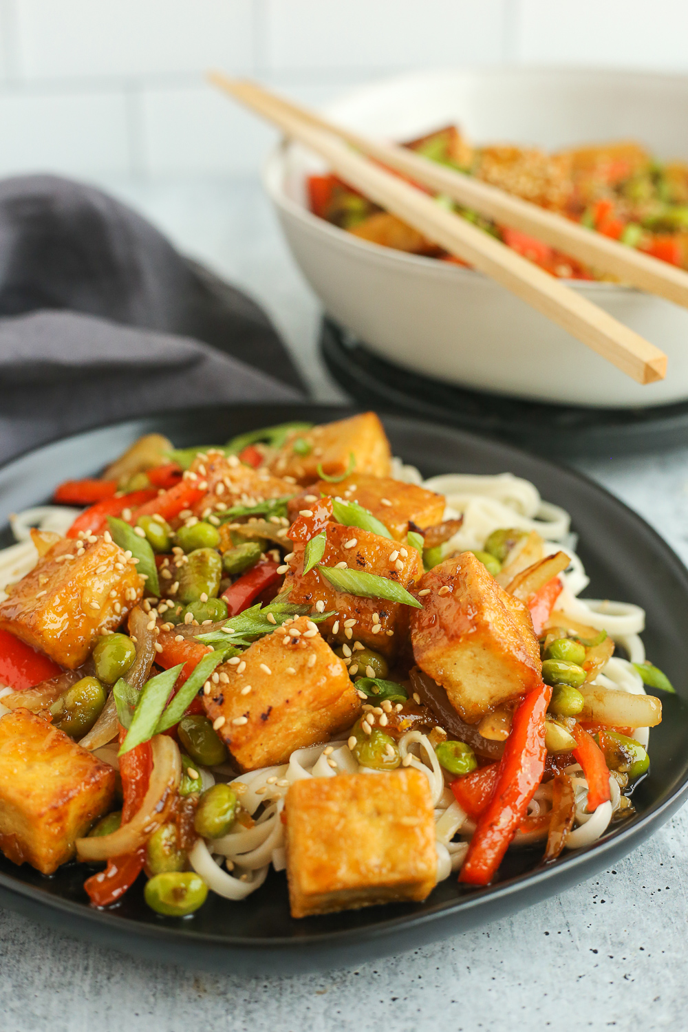 A side view of a serving of Crispy Orange Tofu stir fry on a black ceramic plate, served on top of lo mein noodles with shelled edamame, sliced onions, sliced red bell peppers, and sliced green onions with a toasted sesame seed garnish