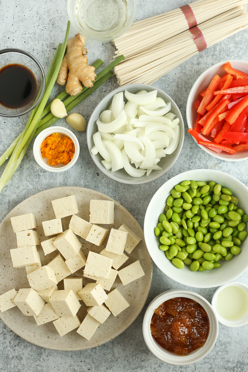 Assortment of ingredients displayed on a grey stone countertop, including a bundle of noodles. ginger, garlic cloves, green onions, soy sauce, sliced onions and red bell pepper, shelled edamame, orange zest and orange marmalade, and a block of firm tofu cut into large cubes