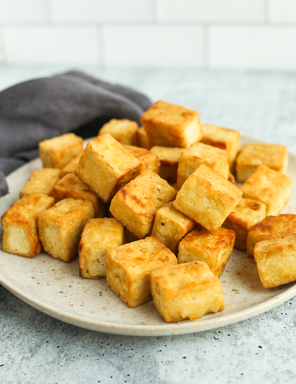 A close up shot of crispy pan fried tofu on a light brown ceramic plate on a grey kitchen countertop