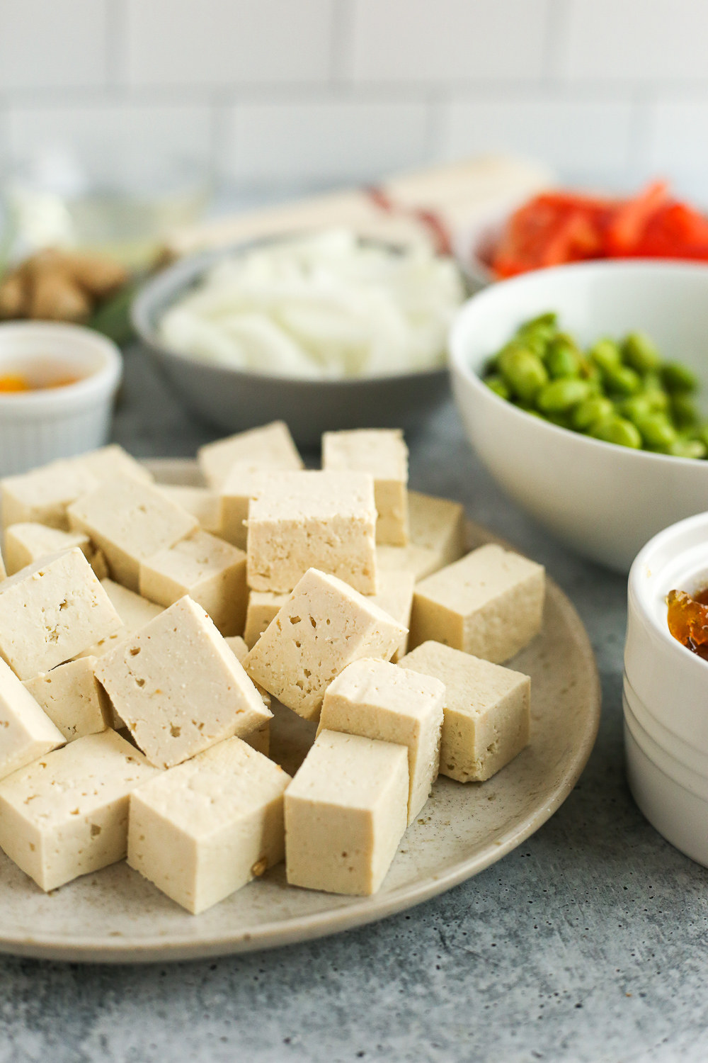 Cubes of firm tofu on a light brown plate with other chopped and prepared vegetables in ramekins of varying sizes in the background