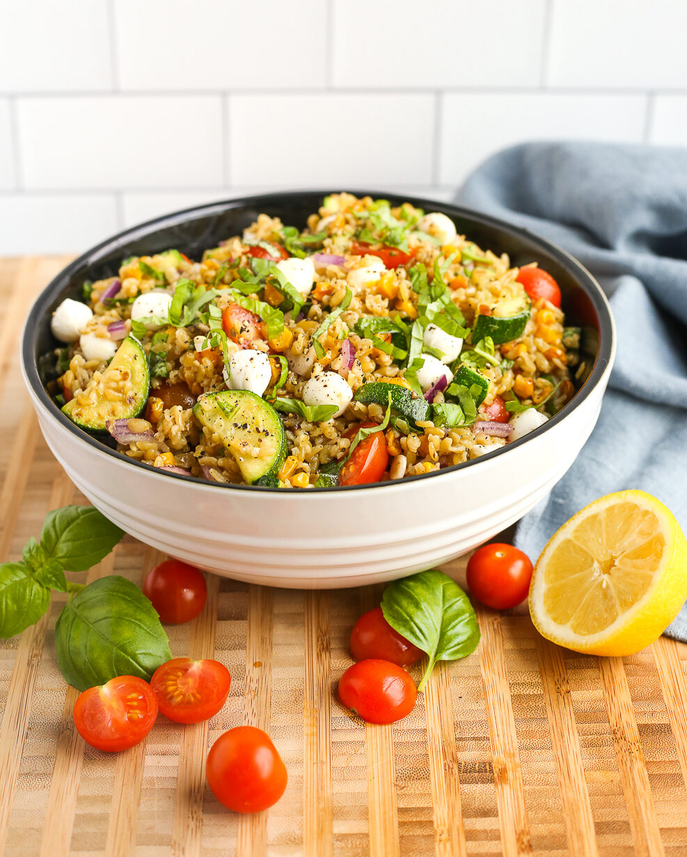 A large mixing bowl on a butcher block countertop filled with a freekeh salad made with cooked freekeh, sliced zucchini, cherry tomatoes, diced red onion, fresh basil, roasted sweet corn, and mozzarella
