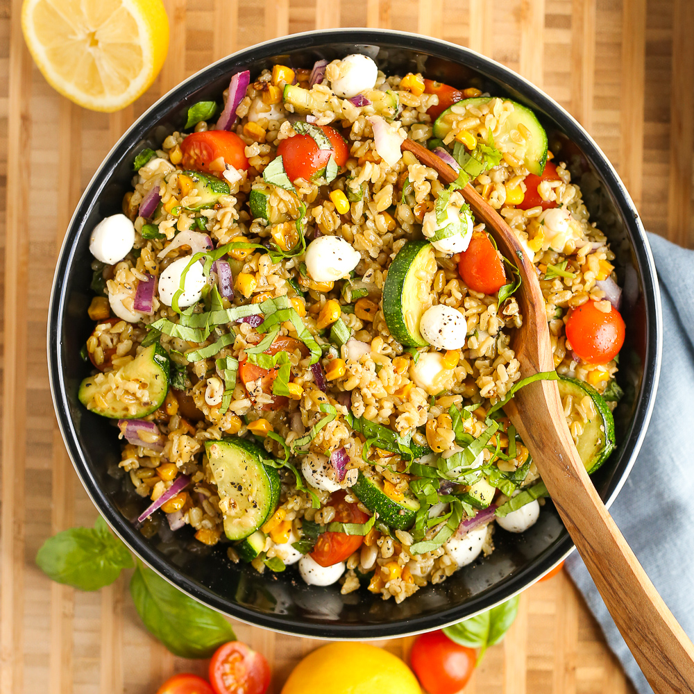 A large mixing bowl sits on a butcher block countertop, filled with a freekeh salad recipe with a wooden spoon resting on the side of the bowl