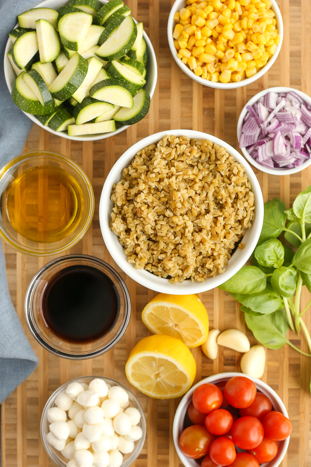 Overhead view of ingredients for summer salad recipes on a butcher block counter, including cooked Freekeh, sliced zucchini, sweet corn, tomatoes, lemons, mozzarella, and garlic cloves