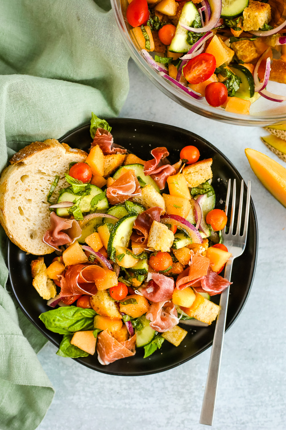 Overhead view of the styled Summer Panzanella Salad with Cantaloupe and Prosciutto recipe, served on a black ceramic plate with a silver fork
