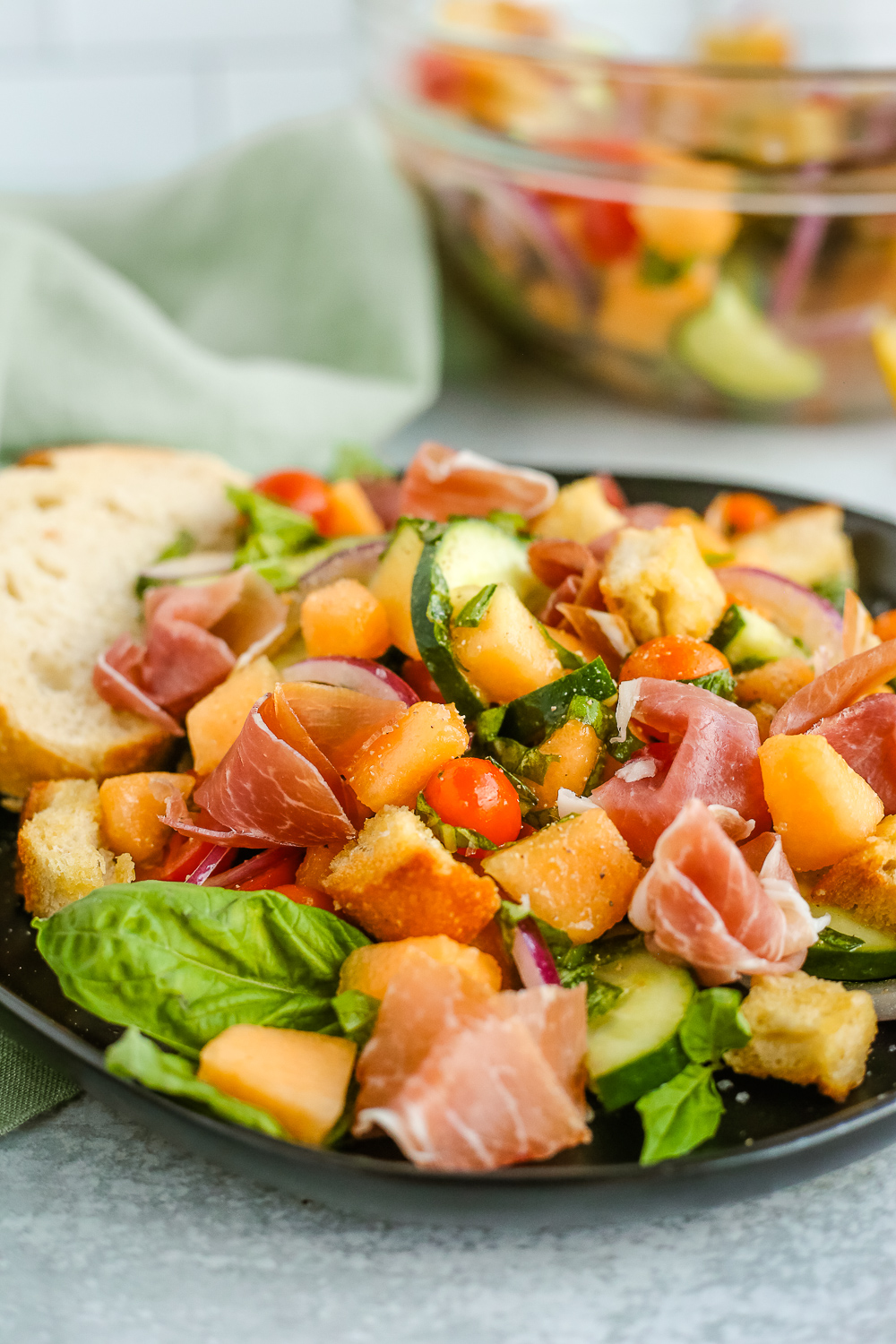 A side view of a summer panzanella salad recipe with tomatoes, cucumbers, onions, cantaeloupe, prosciutto, basil, and bread on a black plate with a clear glass mixing bowl in the background