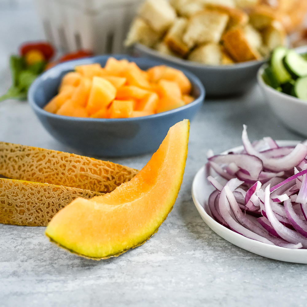 A closer view of a slice of bright orange cantaloupe with the rind, arranged next to the other ingredients for a summer panzanella salad recipe