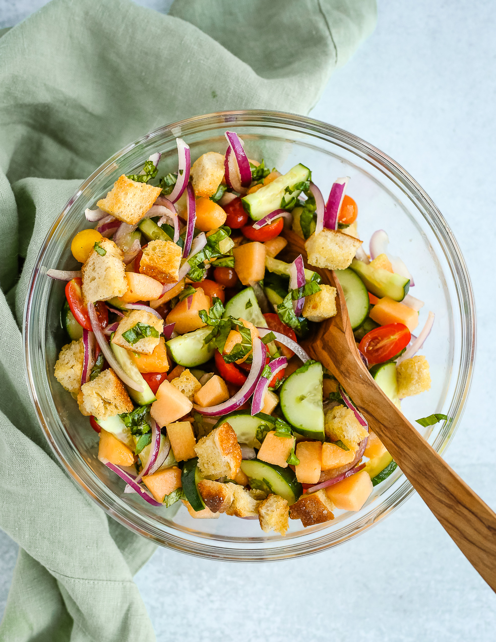 Overhead view into a large clear glass mixing bowl containing a panzanella salad recipe with vibrant, colorful chunks of cantaloupe, cucumber, tomatoes, prosciutto, sliced red onion, and crusty chunks of bread. A wooden serving spoon rests on the side of the bowl