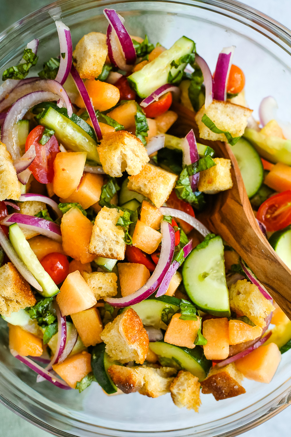 A clear glass mixing bowl with cucumbers, onions, tomatoes, bread, basil, and cantaloupe, mixed together with a wooden serving spoon