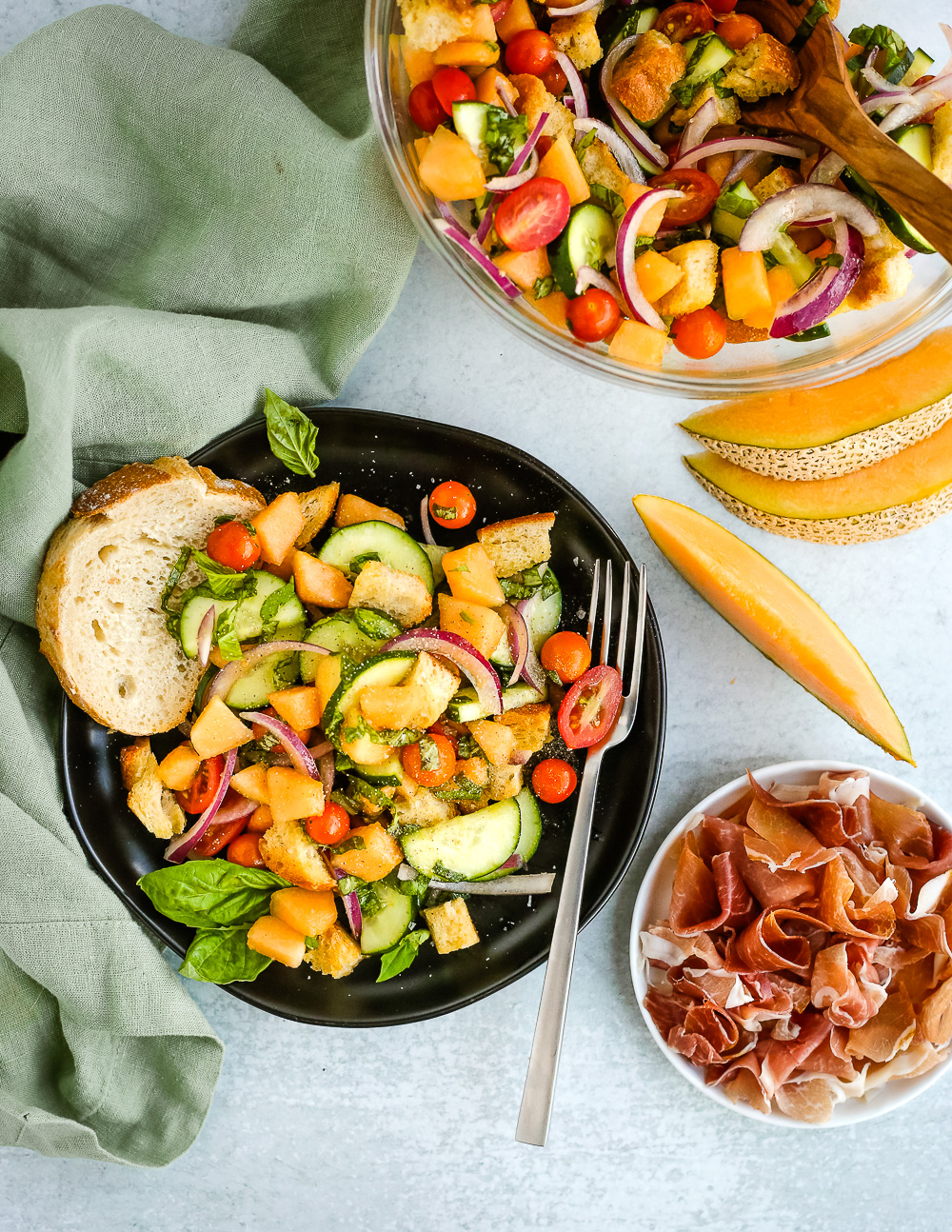 Overhead view of a summer panzanella salad with cantaloupe with prosciutto on a black plate, surrounded by ingredients and a light green linen on a light grey background