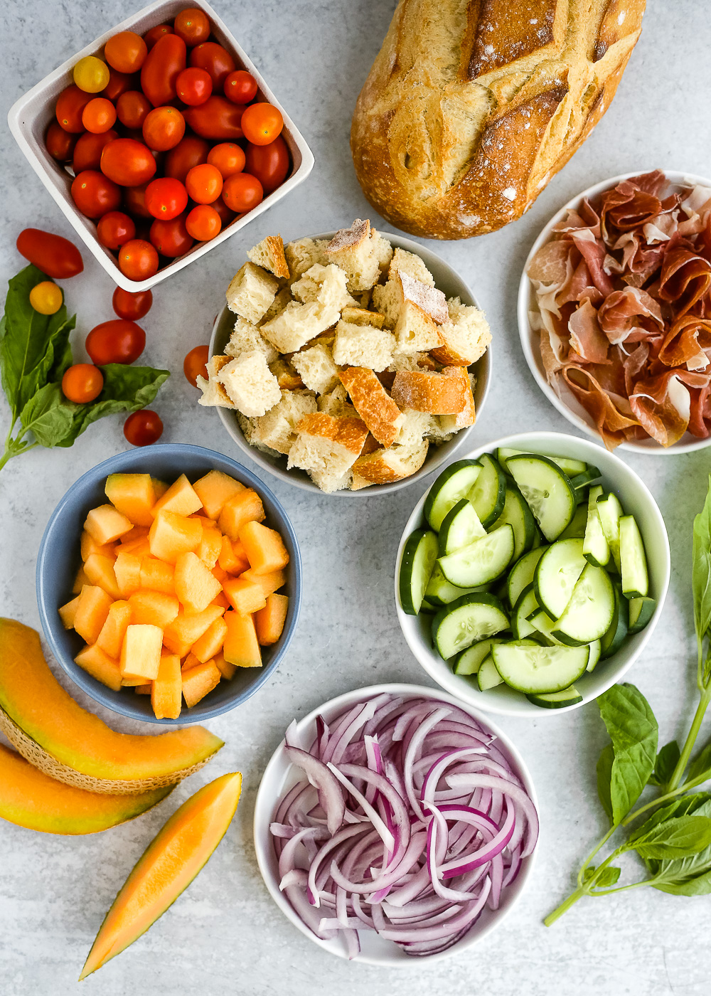 An overhead view of all the ingredients for a summer panzanella salad recipe on a light grey background, including a loaf of Italian bread, tomatoes, prosciutto, basil, cucumbers, cantaloupe, and red onion