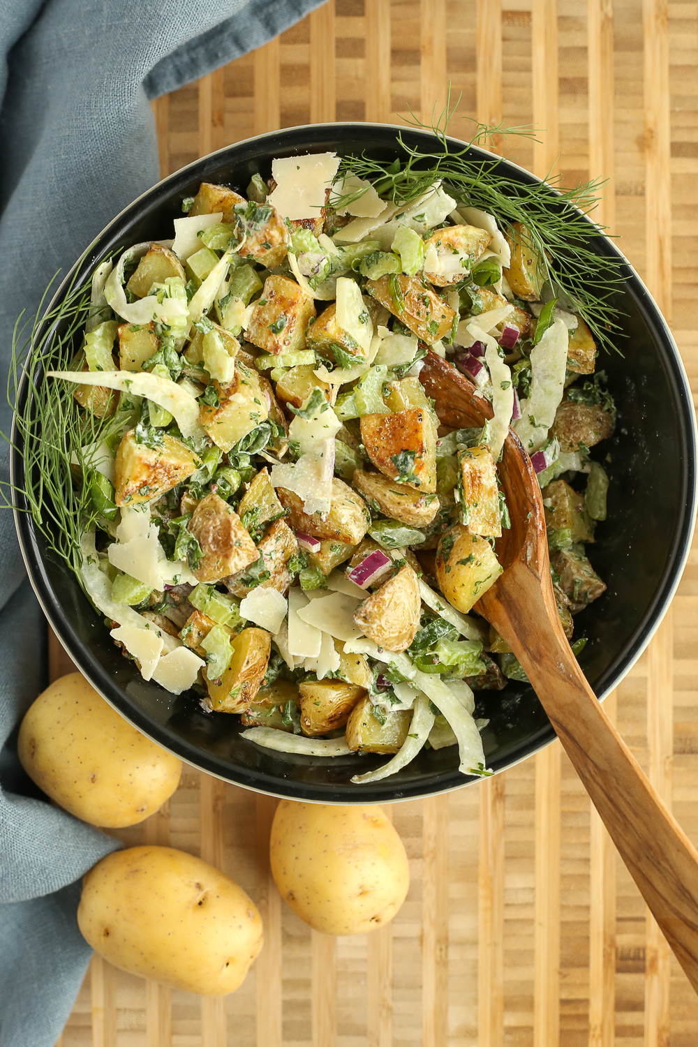 Overhead view of a creamy potato salad in a black ceramic serving bowl, garnished with fennel leaves and parmesan cheese, with a wooden spoon resting on the side of the bowl