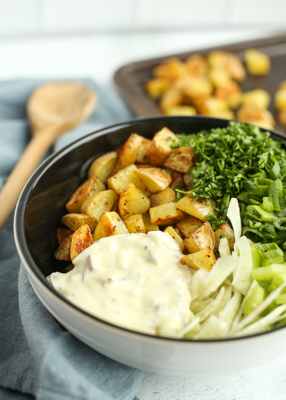 Unmixed ingredients for a creamy potato salad recipe in a white mixing bowl. A sheet pan of roasted potatoes, a wooden spoon, and a blue linen are in the background