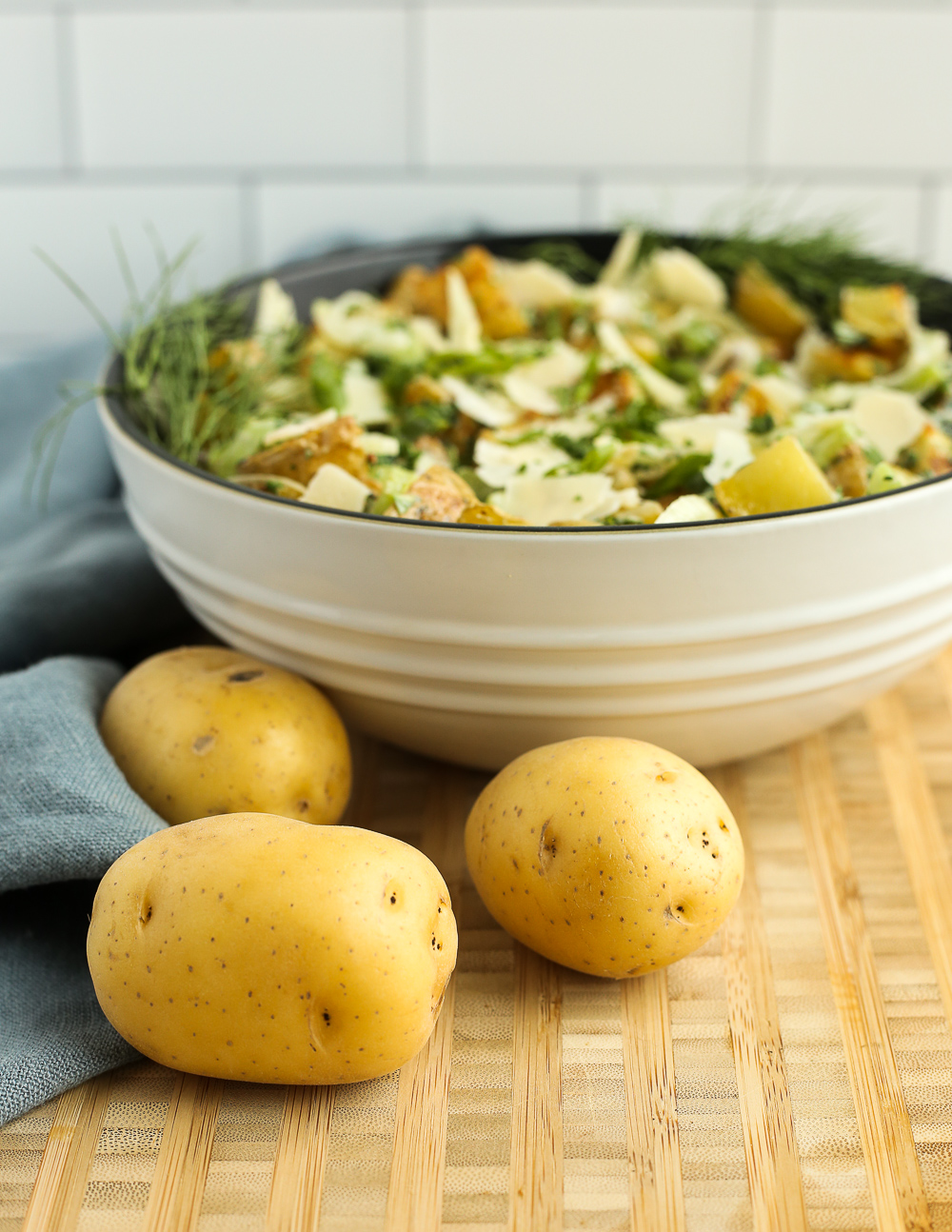 A side view of a white mixing bowl with a creamy potato salad topped with herbs. Three uncooked potatoes are on the butcher block counter in front of the bowl