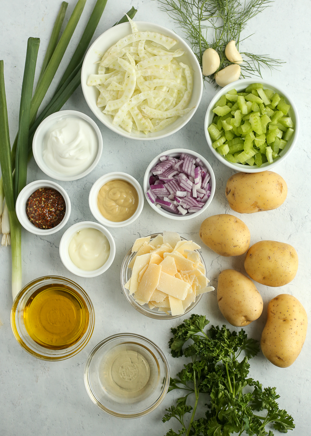 An overhead view of the ingredients of a creamy Greek yogurt potato salad recipe in small white ramekins on a light grey background