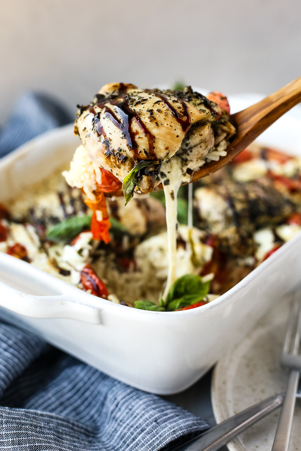 A wooden spoon dishes caprese chicken and rice bake from a while casserole dish, with melted mozzarella cheese stretching from the spoon to the dish
