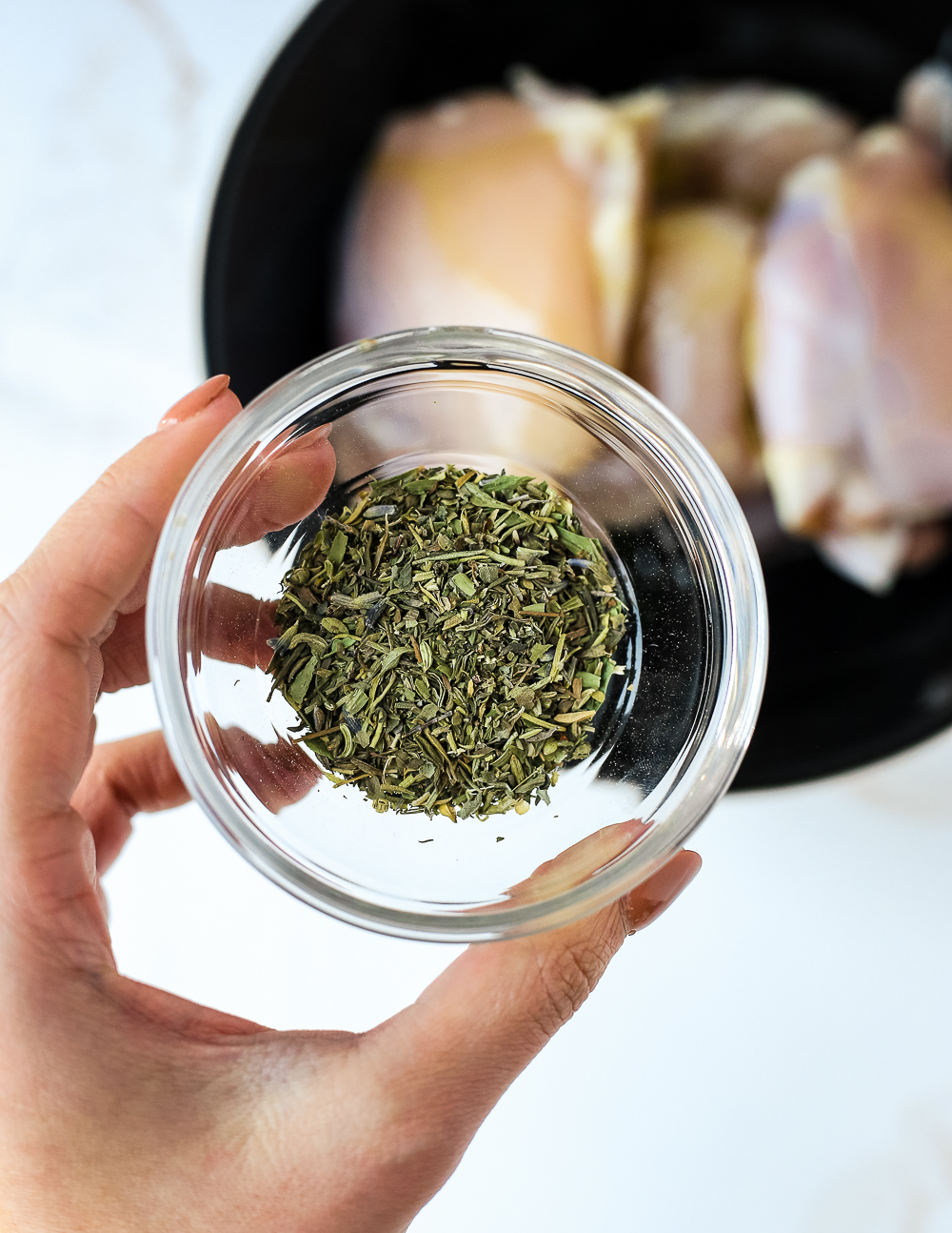 A woman's hand holds a clear glass ramekin of herbs de Provence over a mixing bowl that holds uncooked boneless, skinless chicken thighs