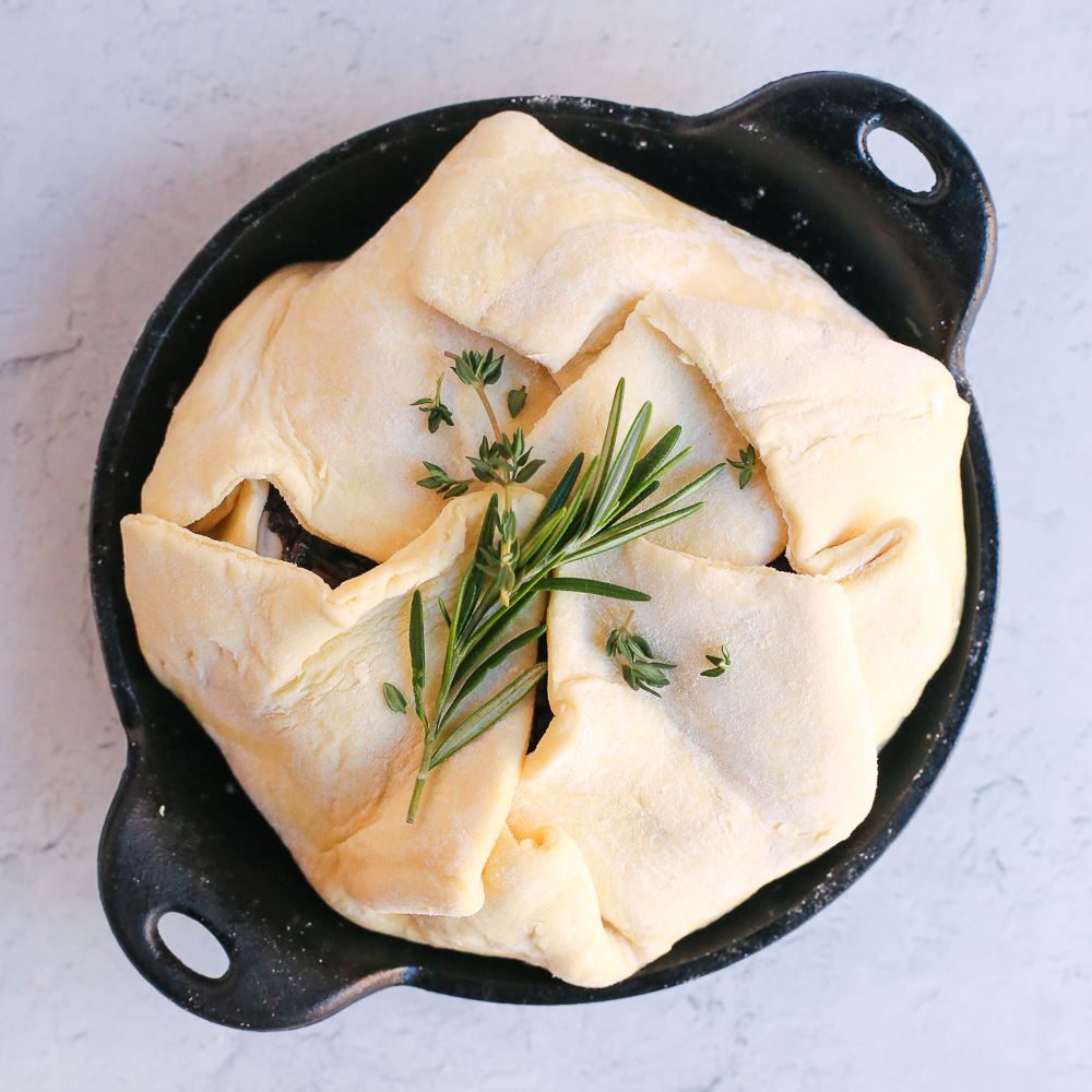 A folded, but unbaked, sheet of puff pastry enclosing a wheel of brie with fresh herbs on top.
