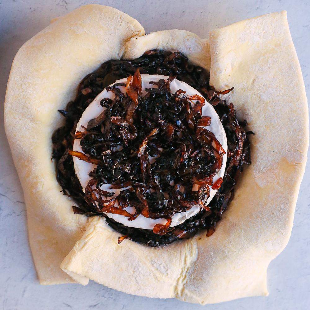A process shot to make a baked brie appetizer, showing an open sheet of puff pastry with a wheel of brie in the center, topped with caramelized onions.