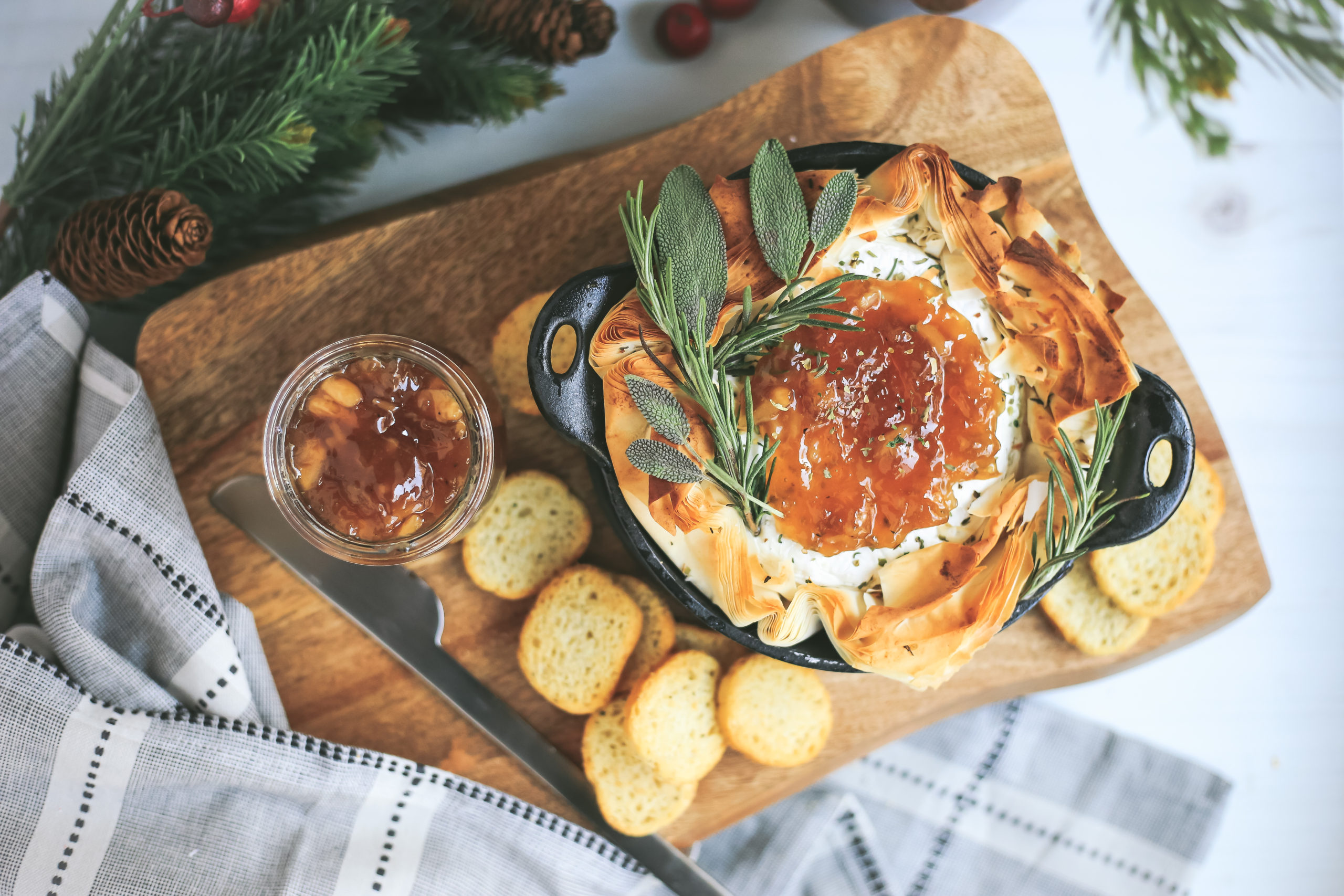 Overhead view of baked brie in phyllo dough served on a wooden platter with crackers and fig jam, garnished with fresh herbs