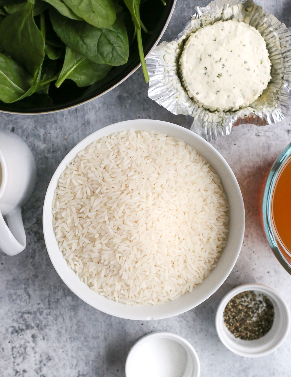 Closeup view of uncooked white rice in a white ceramic bowl on a grey stone countertop, surrounded by additional ingredients