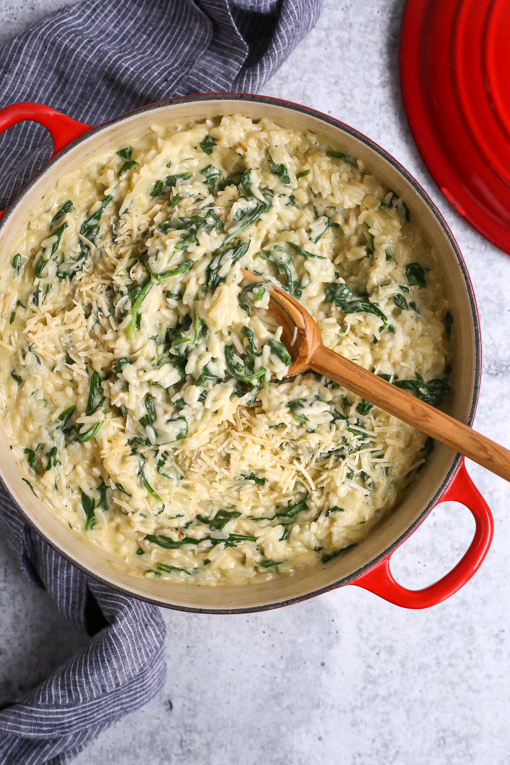 A pot full of cheesy spinach rice with a wooden serving spoon and blue striped linen on a cement kitchen countertop