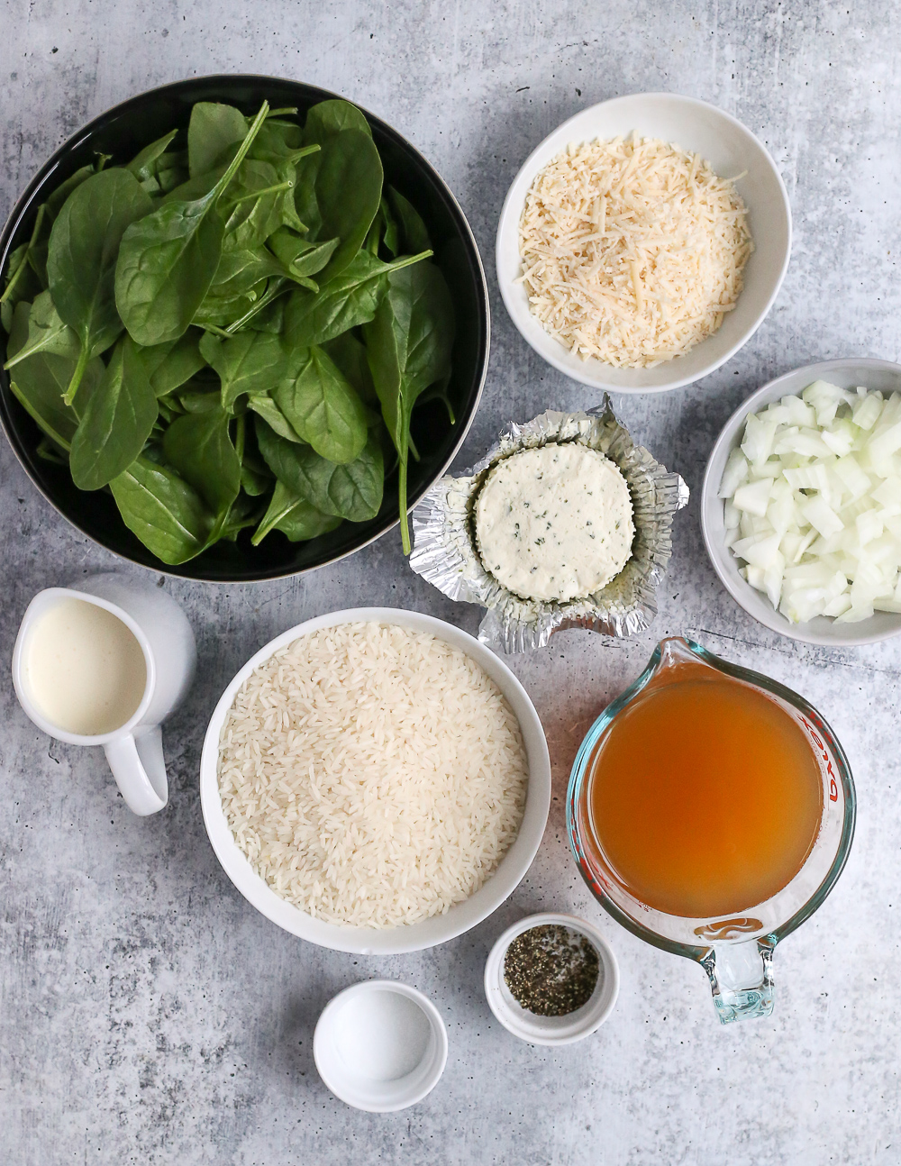 Collection of ingredients for the creamy spinach rice recipe, including uncooked white rice, parmesan cheese, Boursin cheese, fresh spinach, diced onions, heavy cream, vegetable stock, and salt and pepper