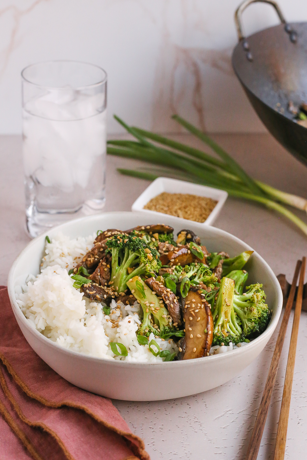 Beef and Broccoli with Mushrooms served in a bowl with white rice, garnished with green onions and sesame seeds, served with chopsticks, a glass of water, and a light pink napkin in a kitchen scene