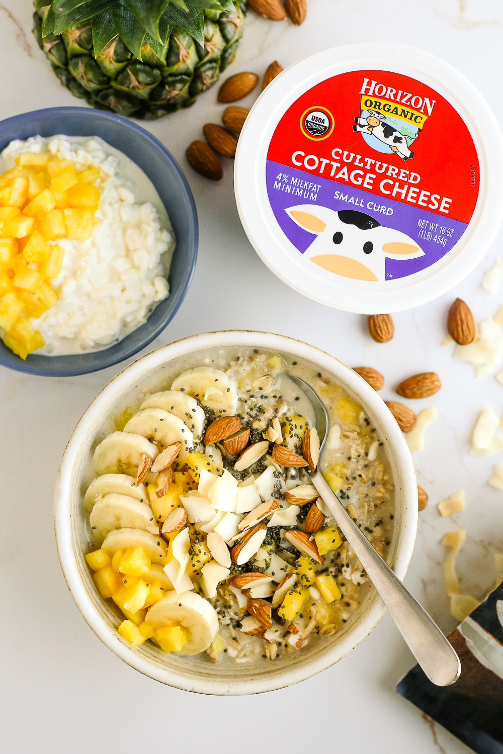 Overhead view of cottage cheese in oatmeal, showing a container of cottage cheese, a bowl of cottage cheese and pineapple, and an oatmeal bowl with bananas, pineapple, almonds, and coconut