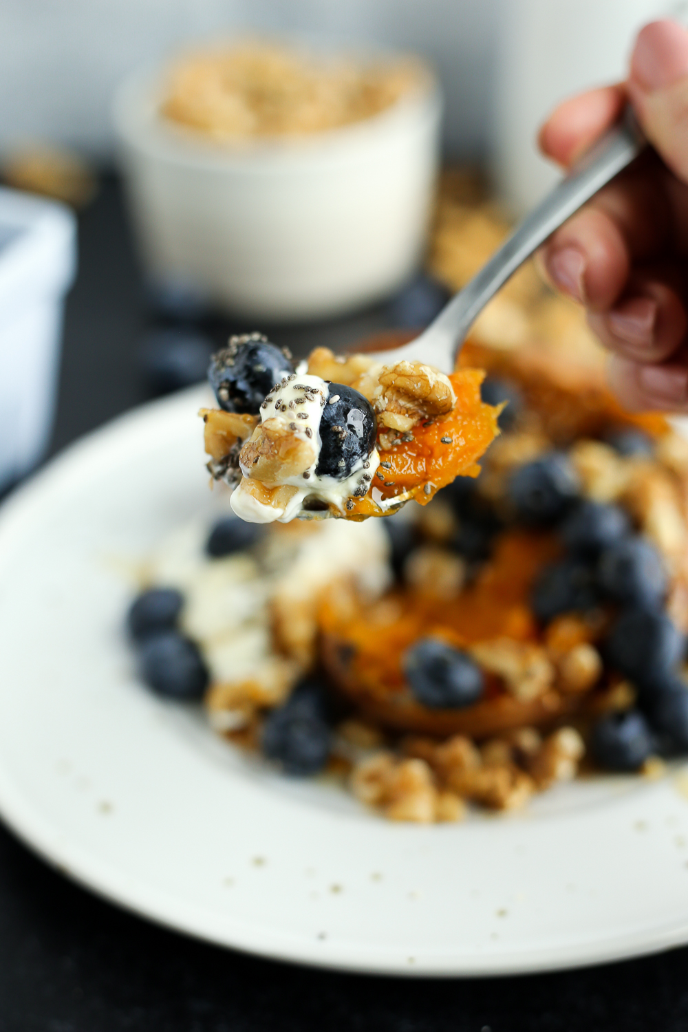 A woman's hand holds a silver spoon piled high with cooked sweet potato, a few blueberries, chopped walnuts, and a small amount of yogurt. The mixed bite is topped with a few chia seeds and the rest of the dish is blurred in the background