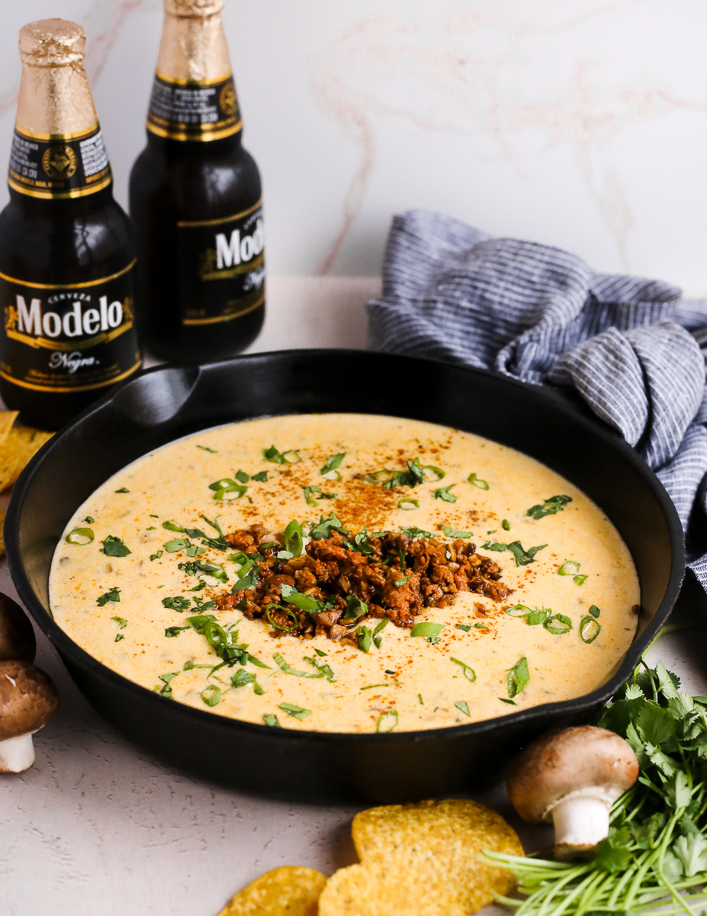 Styled image of a green chile queso recipe, served in a black cast iron skillet on a kitchen counter with tortilla chips and bottles of Mexican beer