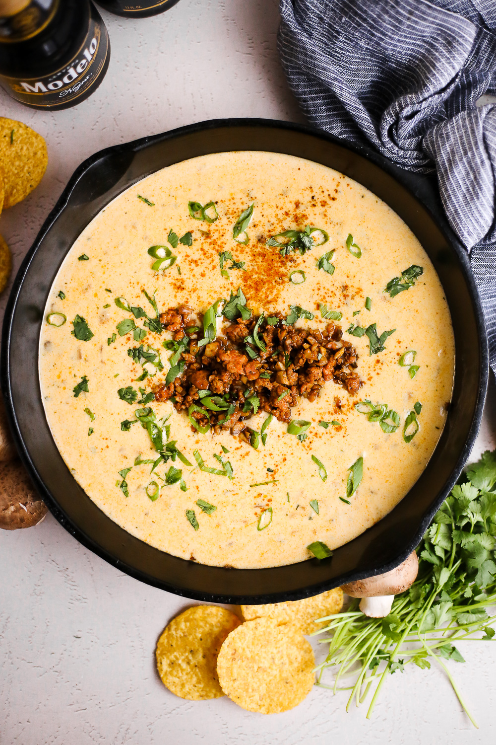Overhead view of green chile queso in a black cast iron skillet, garnished with cilantro, green onions, chorizo, and mushrooms, served with tortilla chips and bottles of beer
